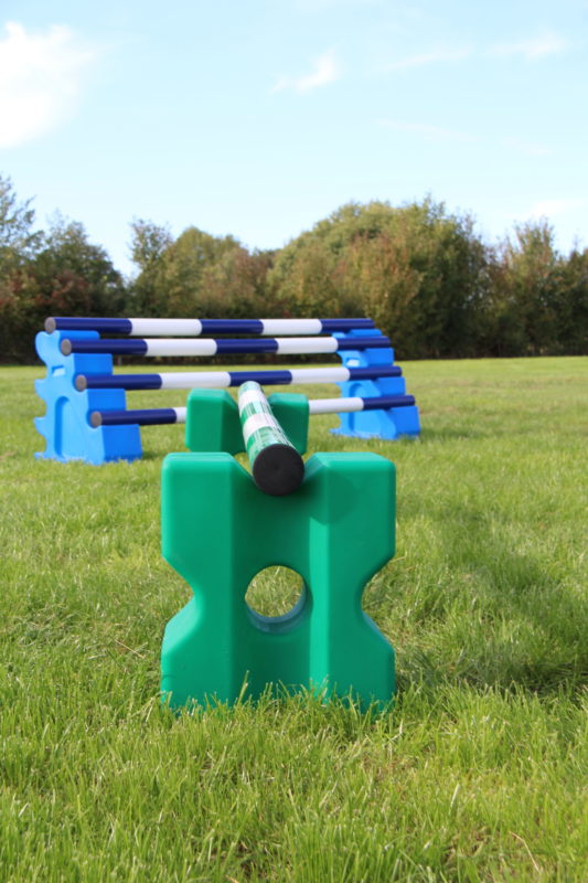 Das passende Trainingszubehör optimiert das Training. Trainingszubehör auf einer Wiese: Im Vordergrund ein grünes Cavaletti "Cona", im Hintergrund ein blauer Sprung bestehend aus den Horseblocks und 4 Stangen.